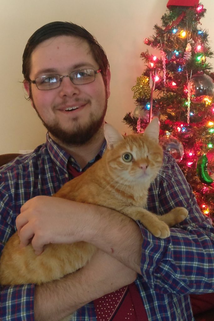 Photograph of Simon Miller, a white man with a beard and glasses holding a one eyes cat in front of a christmas tree